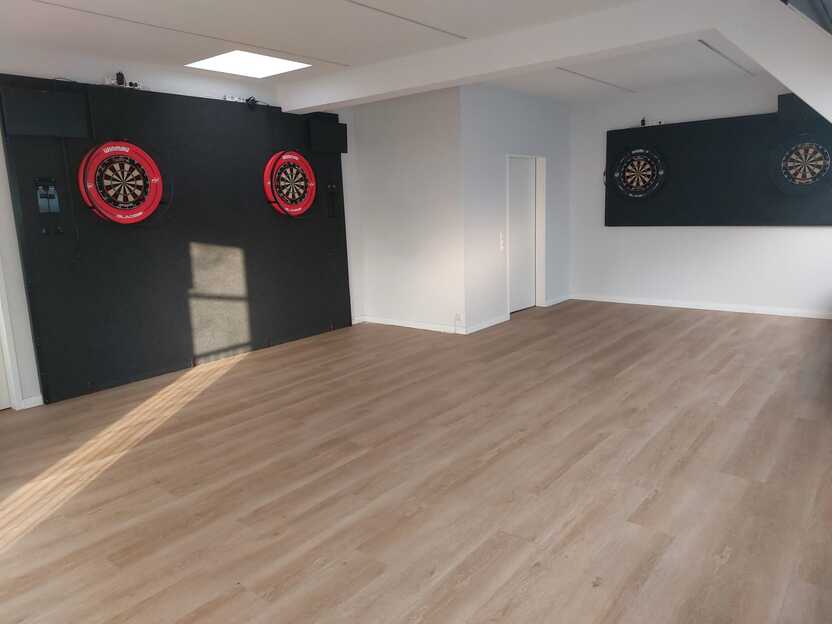 Leere Darts-Spielhalle mit zwei Dartboards an schwarzen Wänden und einem hellen Holzboden.