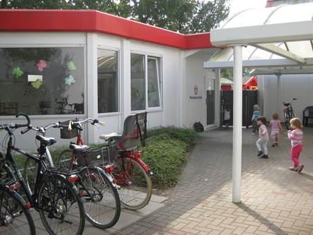 Eingang eines Kindergartens mit Fahrrädern im Vordergrund und spielenden Kindern auf dem Weg zur Tür.