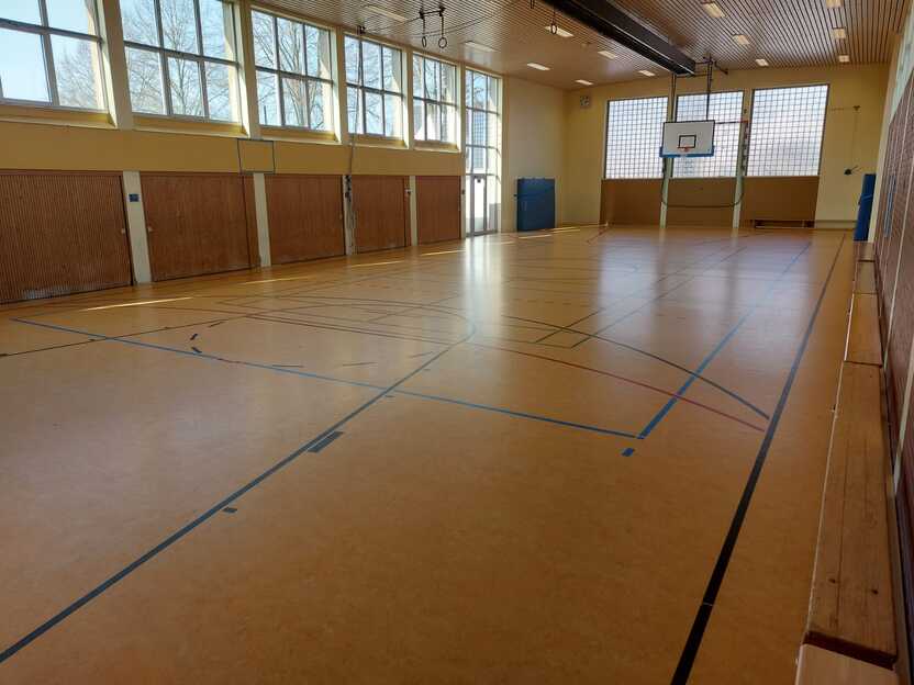 Leerer Sporthalle mit Holzfu&szlig;boden, gro&szlig;en Fenstern und Basketballkorb an der Wand. Licht scheint durch die Fenster.