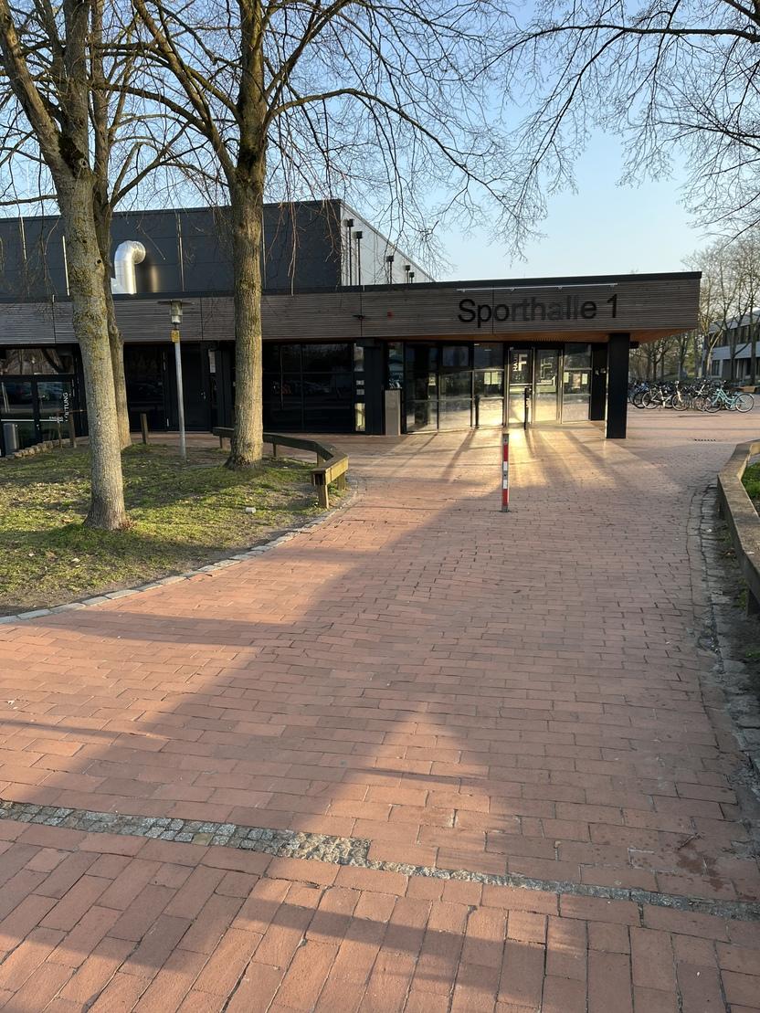 Eingang zur Sporthalle 1 mit gepflastertem Weg und B&auml;umen, im Hintergrund stehen Fahrr&auml;der.