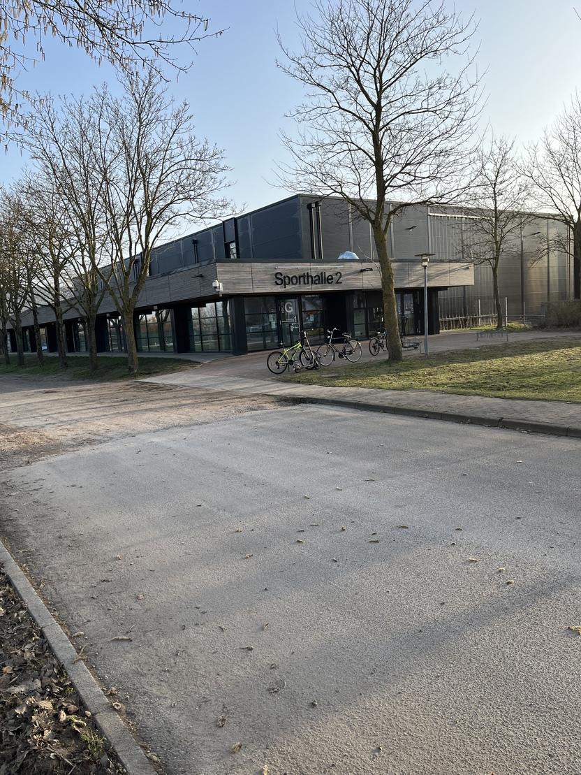 Sporthalle 2 mit Eingang, Radst&auml;nder und B&auml;umen in einer ruhigen Umgebung unter blauem Himmel.
