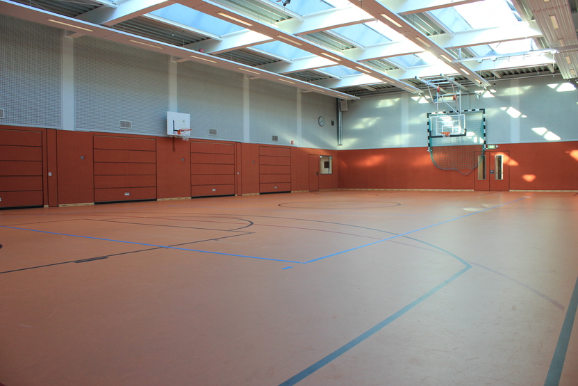 Leere Sporthalle mit oranger&uuml;ckiger Bodenfl&auml;che, Basketballkorb und gro&szlig;en Fenstern.