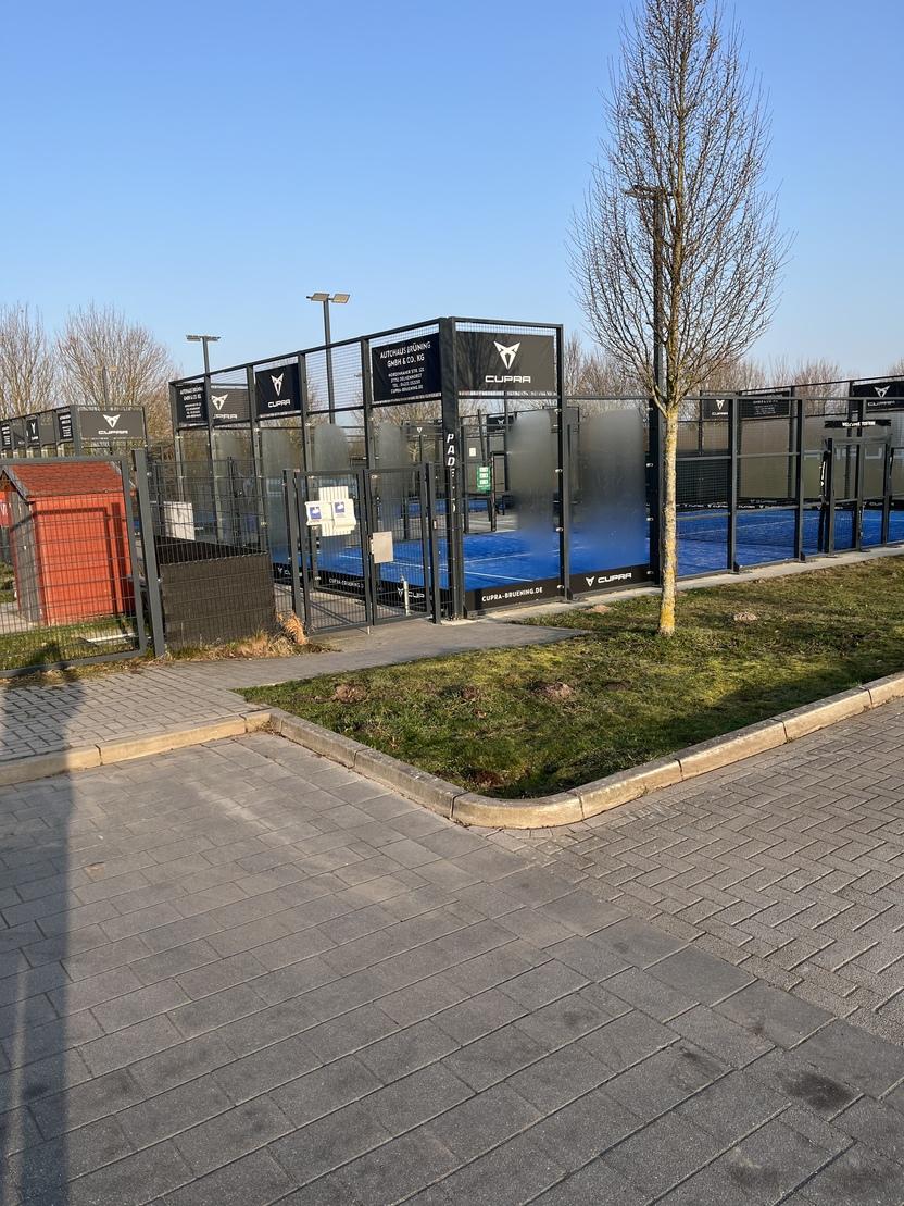 Paddelplatz mit blauer Spielfl&auml;che, umgeben von einem hohen Zaun und Beschilderungen, bei klarem Himmel.