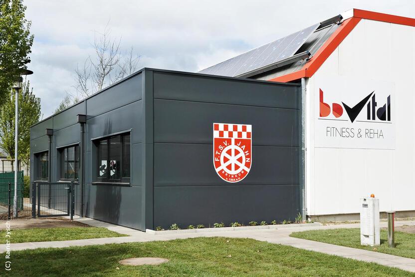 Moderne Sporteinrichtung mit schwarz-grauer Fassade, Vereinswappen und Schild „bovital FITNESS & REHA“.