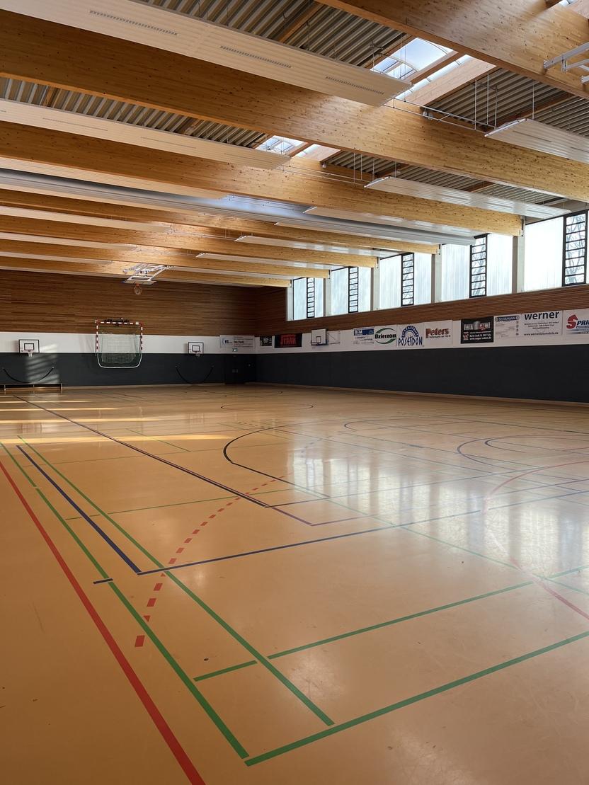 Innenaufnahme einer Sporthalle mit Holzdecke, mehreren Basketballk&ouml;rben und markierten Spielfeldlinien auf dem Boden.
