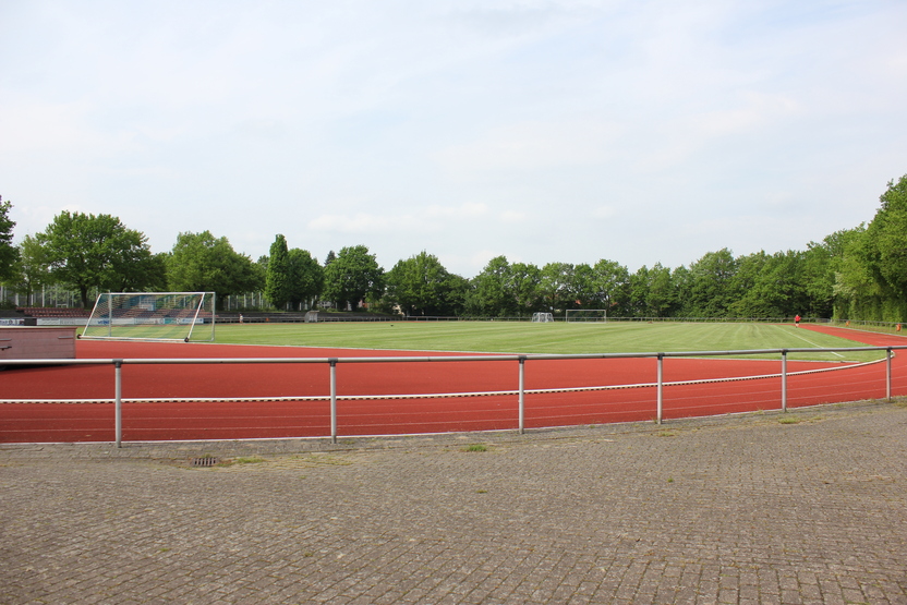 Blick auf einen Sportplatz mit gr&uuml;nem Rasen und Laufbahn, umgeben von B&auml;umen und einem Fu&szlig;balltor.