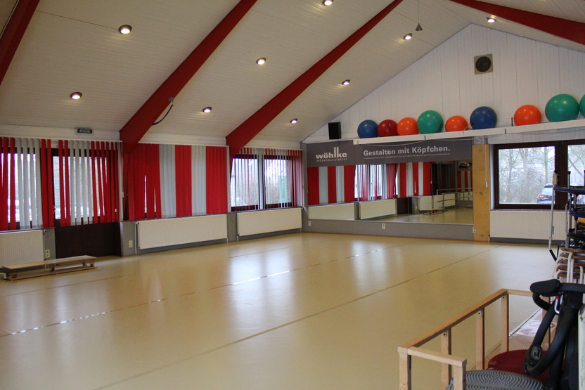 Leerer Raum mit hellen W&auml;nden, gro&szlig;en Fenstern und bunten Fitnessb&auml;llen an der Wand. Spiegel gegen&uuml;ber der Fenster.