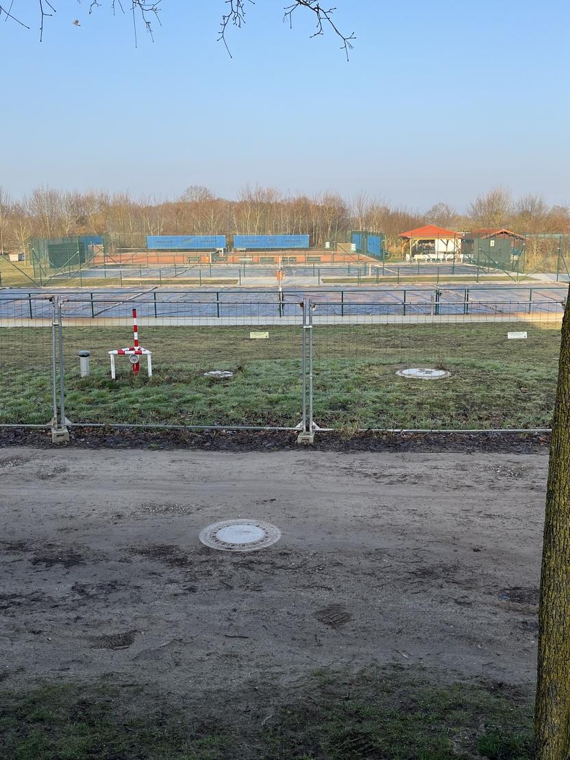 Blick auf einen eingez&auml;unten Tennisplatz mit mehreren Spielfeldmarkierungen und einem &uuml;berdachten Bereich im Hintergrund.