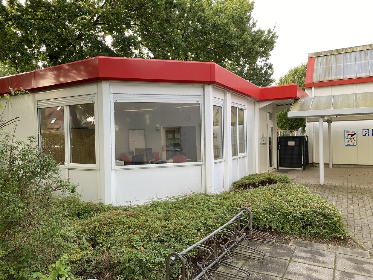 Eingang eines modernen, hellen Geb&auml;udes mit roten Dach und gro&szlig;en Fenstern, umgeben von gr&uuml;ner Bepflanzung.