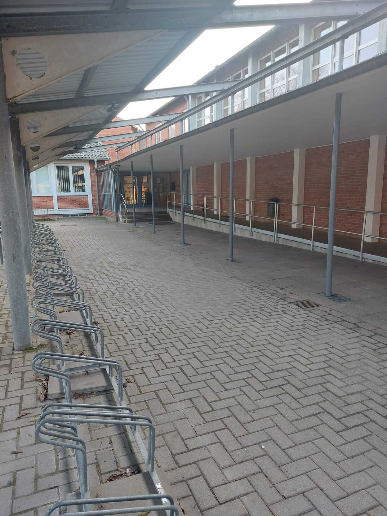 &Uuml;berdachter Gang neben einem Geb&auml;ude, mit mehreren Fahrradst&auml;ndern und gepflastertem Boden. Eingang zum Geb&auml;ude sichtbar.