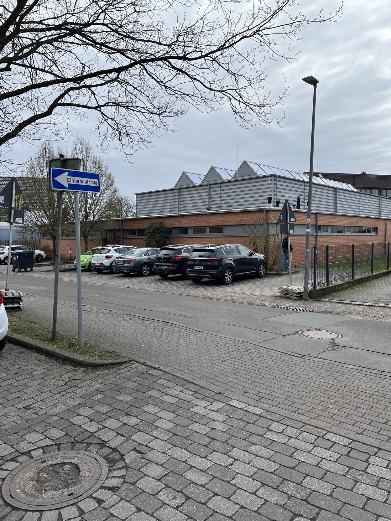 Blick auf ein Parkplatz mit mehreren Autos und einem Wegweiser zur Einbahnstra&szlig;e vor einem modernen, roten Geb&auml;ude.