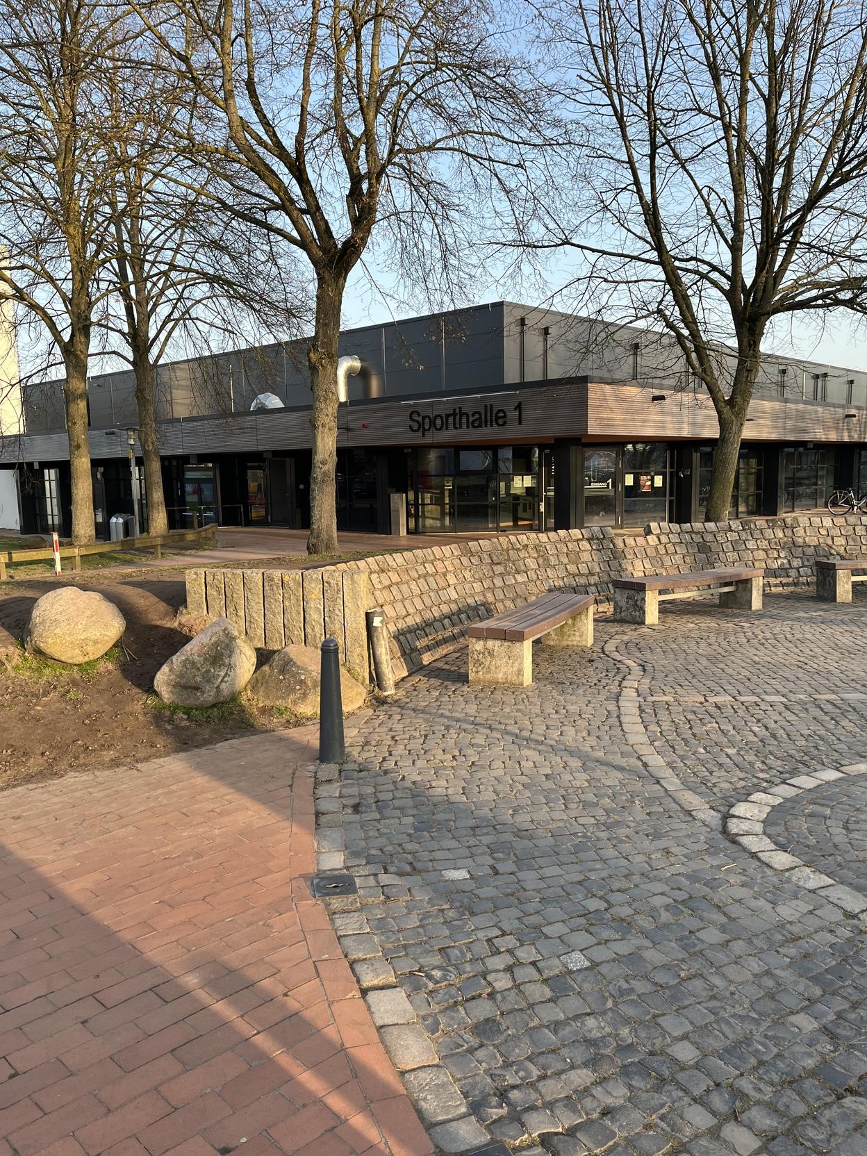 Eingang zur Sporthalle 1, umgeben von B&auml;umen und einer gepflasterten Fl&auml;che mit B&auml;nken und Steinen.