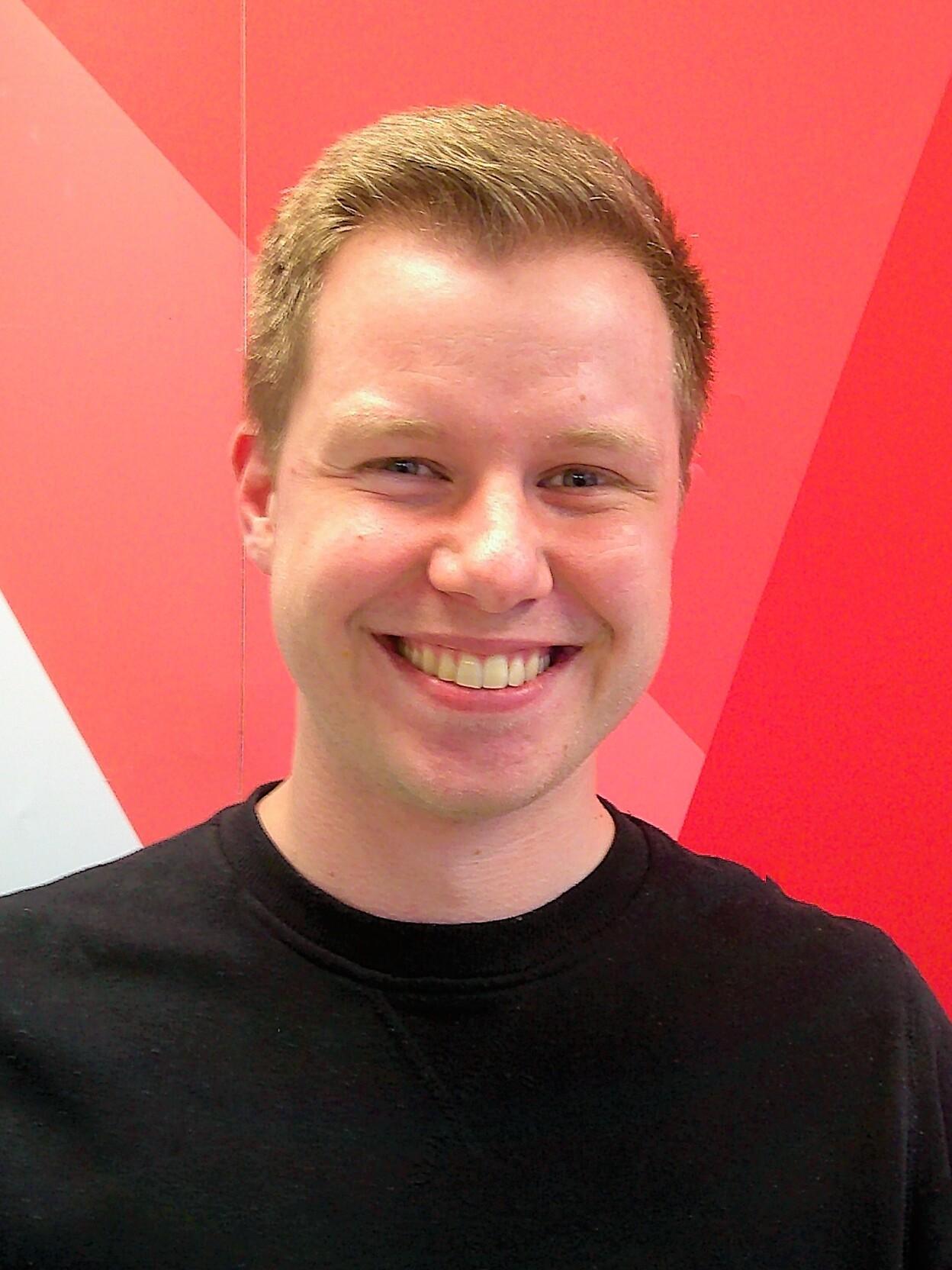 L&auml;chelnder junger Mann mit kurzer Frisur vor einer rot-wei&szlig; grafischen Wand. Tr&auml;gt ein schwarzes Sweatshirt.