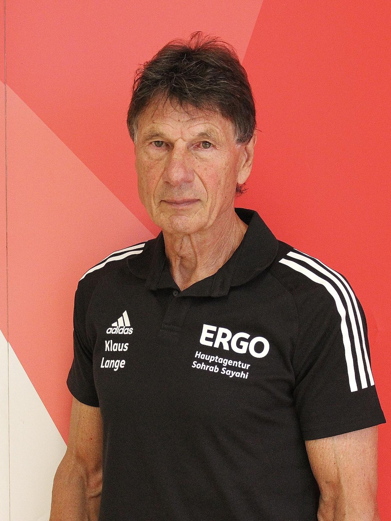 Mann mit dunklem Haar in einem schwarzen Sporthemd steht vor einer rot-wei&szlig;en Wand. Schriftzug auf dem Hemd und Wand sichtbar.