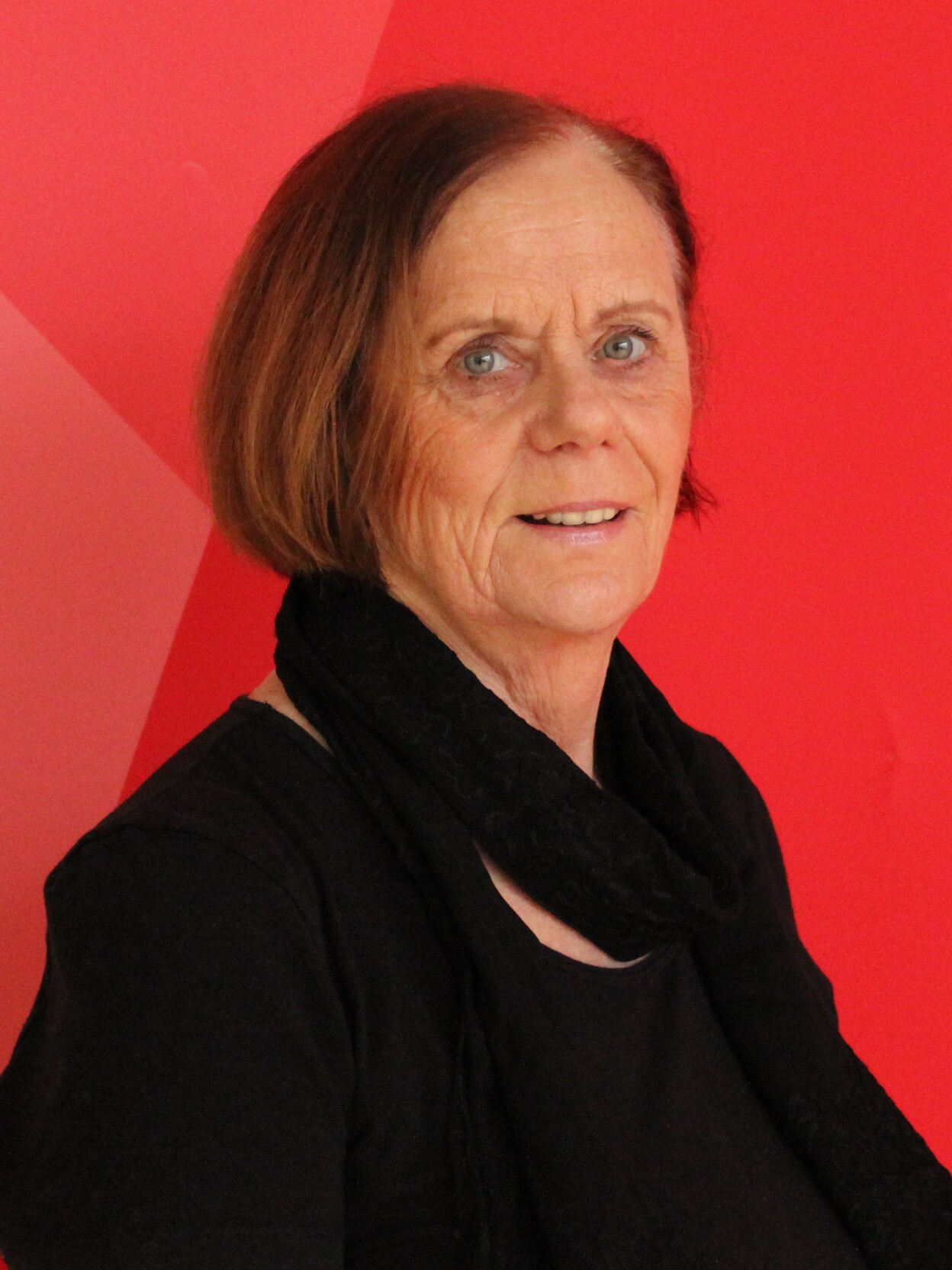 Portr&auml;t einer l&auml;chelnden Frau mit schulterlangen Haaren und schwarzem Oberteil vor einem roten, geometrischen Hintergrund.