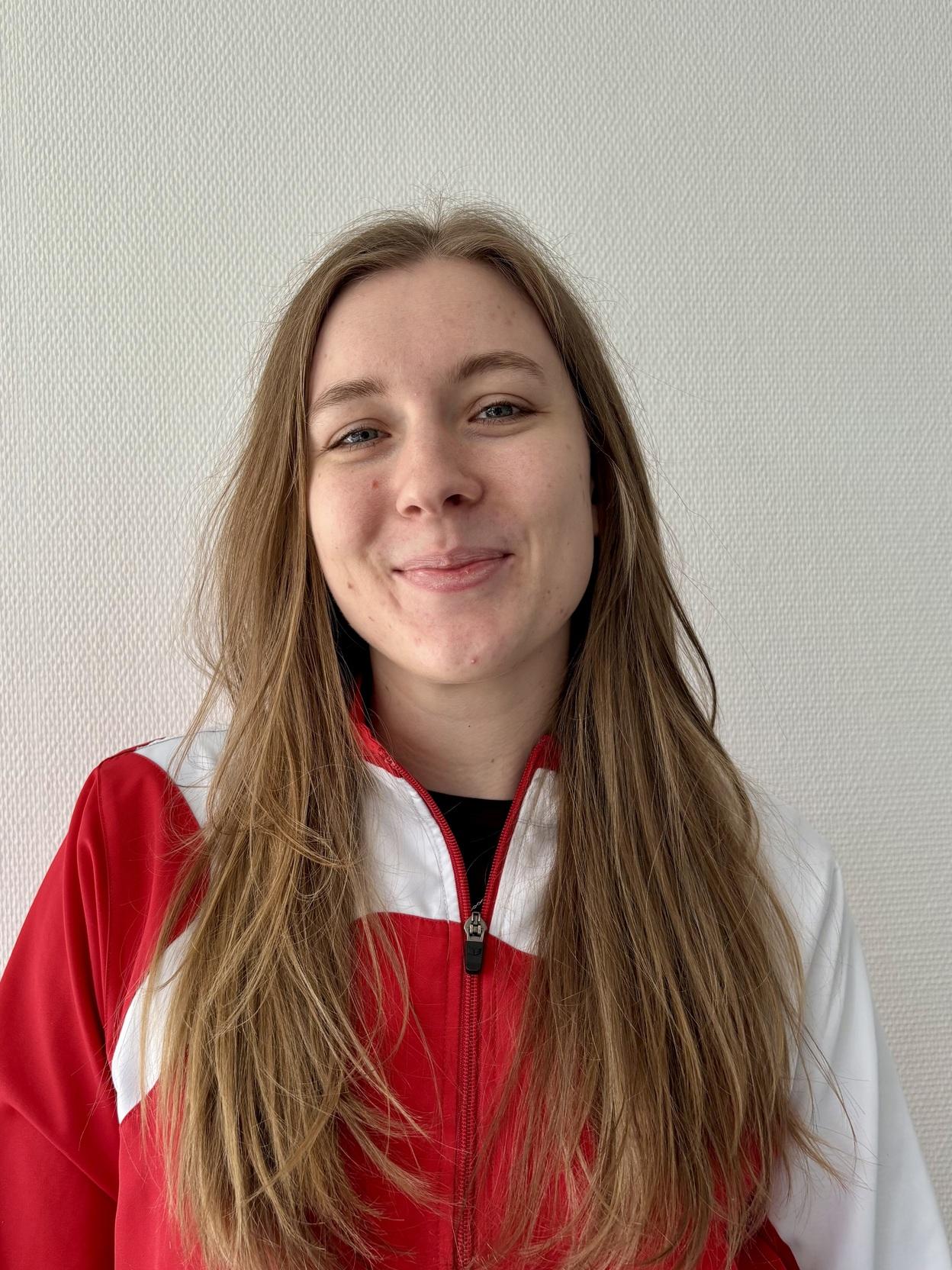 Junge Frau mit langen, blonden Haaren in roter und wei&szlig;er Sportjacke vor hellem Hintergrund.