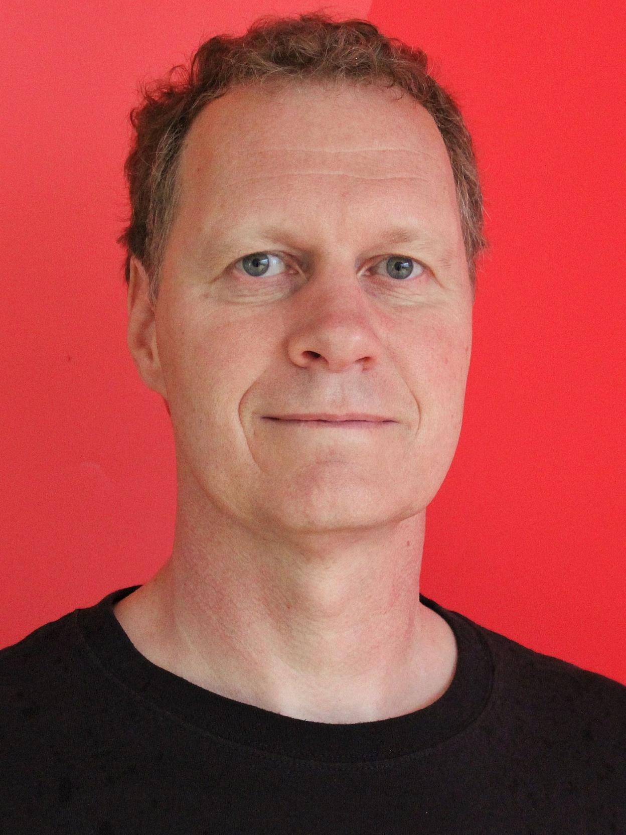 Portr&auml;t eines Mannes mit kurzen, lockigen Haaren vor einem roten Hintergrund, tr&auml;gt ein schwarzes T-Shirt mit Basketballmotiv.