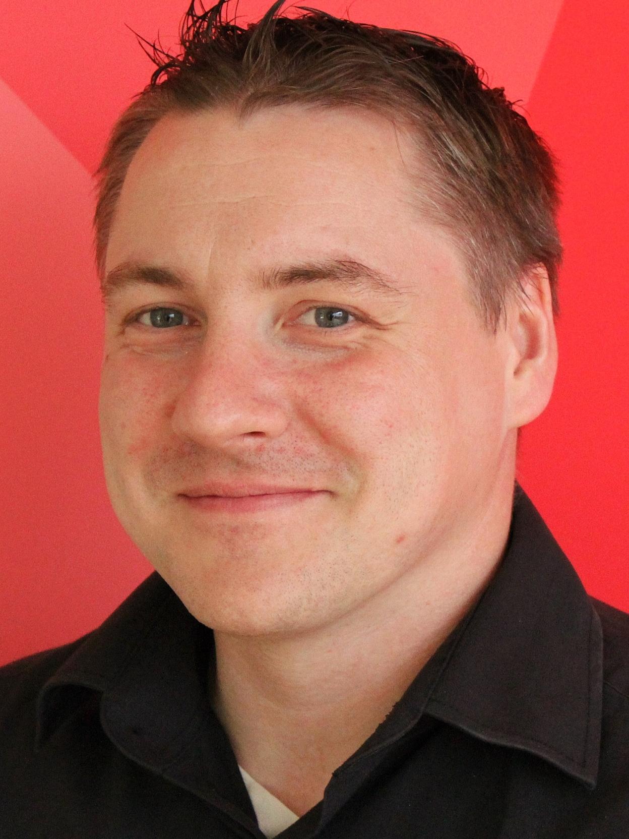 Portr&auml;t eines l&auml;chelnden Mannes mit kurzen, braunen Haaren vor einem roten Hintergrund. Er tr&auml;gt ein schwarzes Hemd.