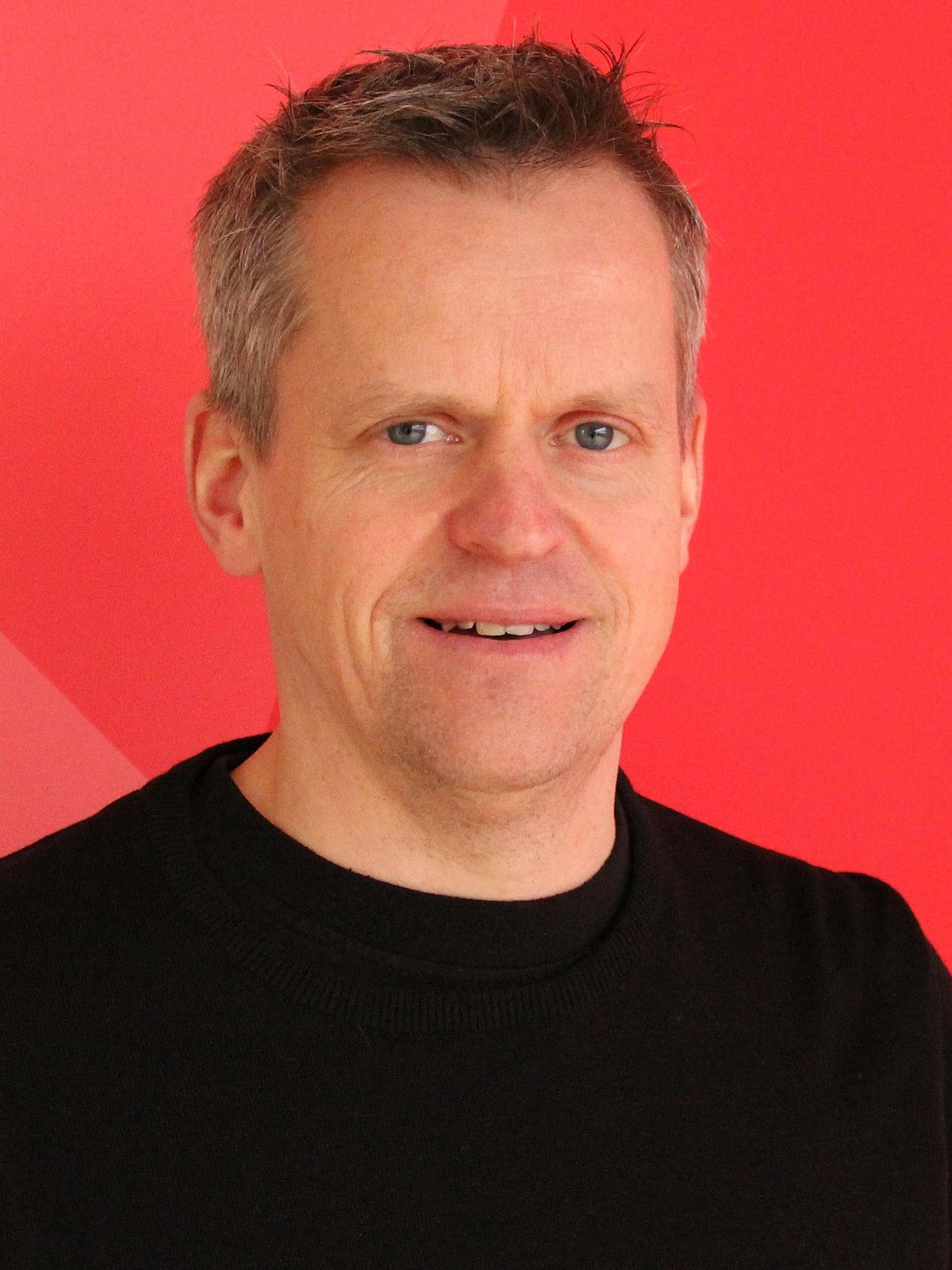 Portr&auml;t eines Mannes mit kurzem grauen Haar, der in einem schwarzen Pullover vor einem roten Hintergrund steht.