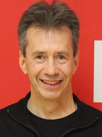 Portrait eines l&auml;chelnden Mannes mit grauen Haaren, der einen schwarzen Trainingsanzug mit rotem Schriftzug tr&auml;gt, vor rotem Hintergrund.