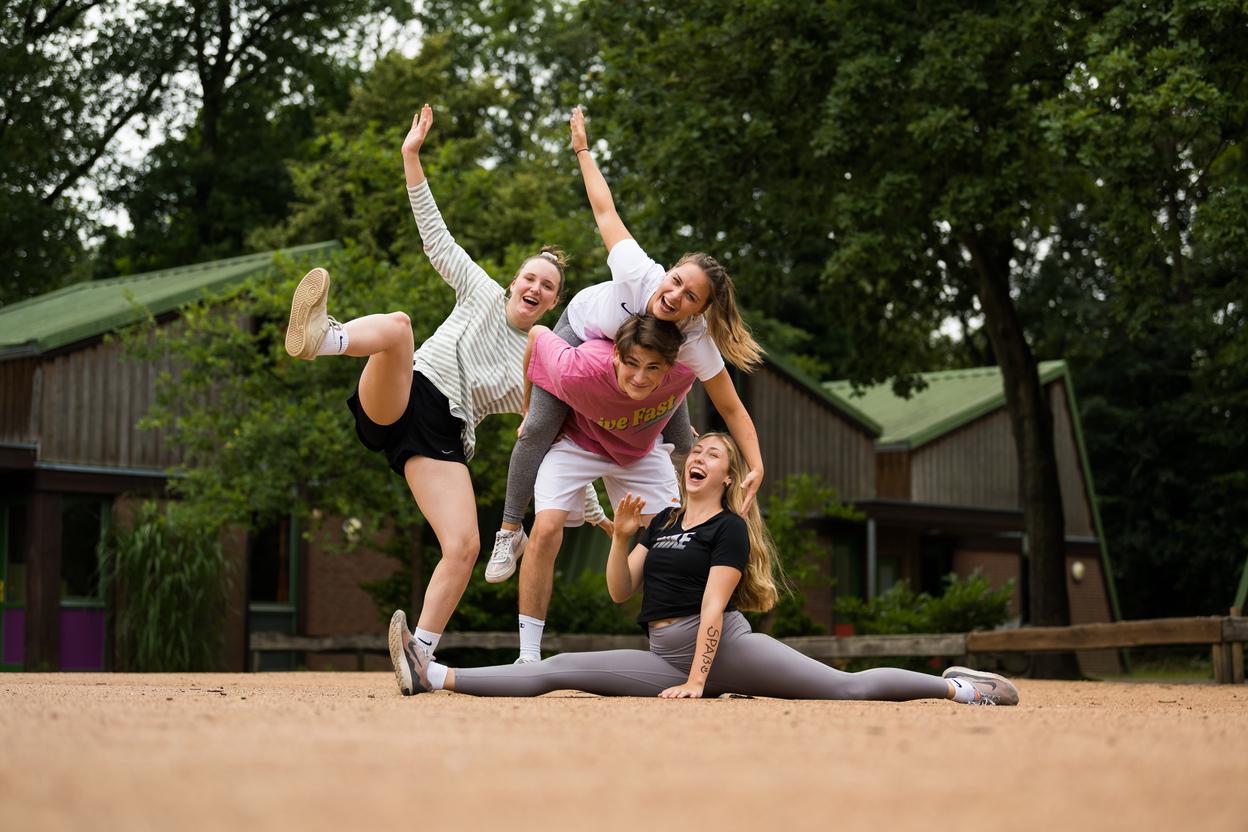 Vier junge Frauen posieren fröhlich auf einer sandigen Fläche, eine sitzt im Spagat, während die anderen sie umarmen.