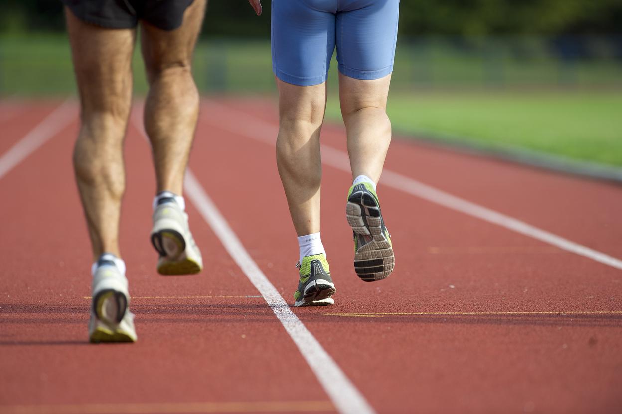 Zwei Läufer, von hinten gesehen, joggen auf einer roten Laufbahn. Der eine trägt kurze Sportshorts, der andere eine blaue Hose.