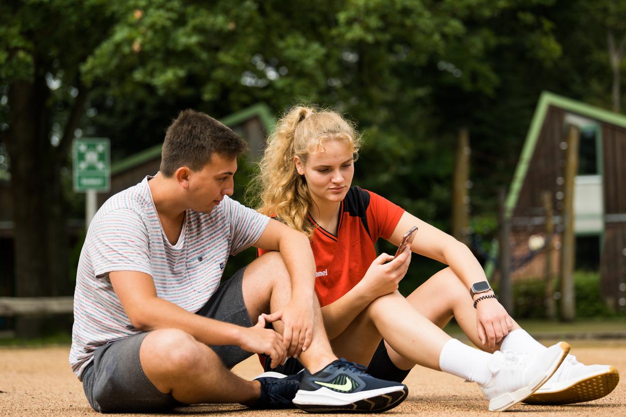 Zwei Jugendliche sitzen im Freien und schauen auf ein Smartphone. Ein Junge in gestreiftem T-Shirt und ein Mädchen in rotem Oberteil.