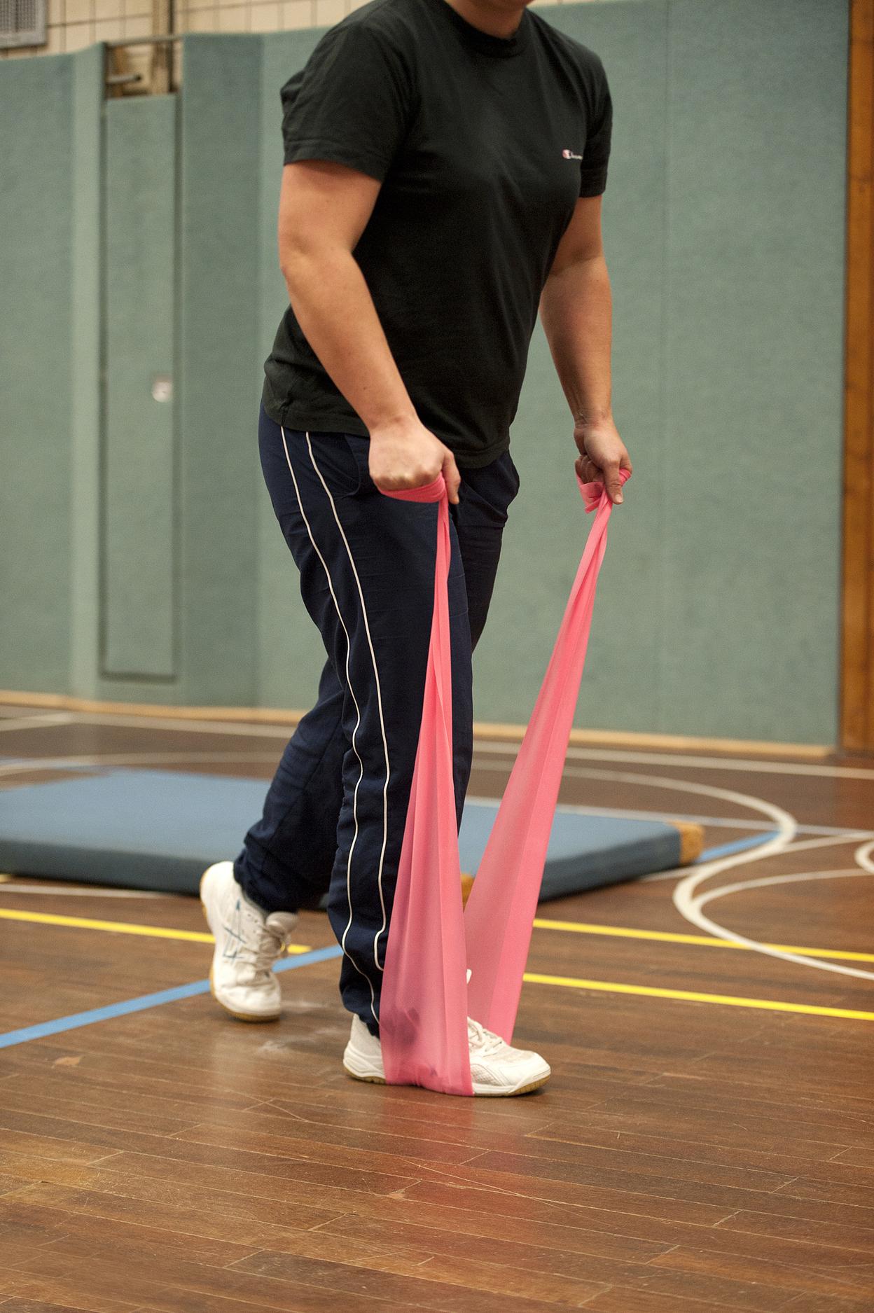 Person in Sportkleidung zieht mit beiden Händen an einem roten Widerstandsband in einer Sporthalle.
