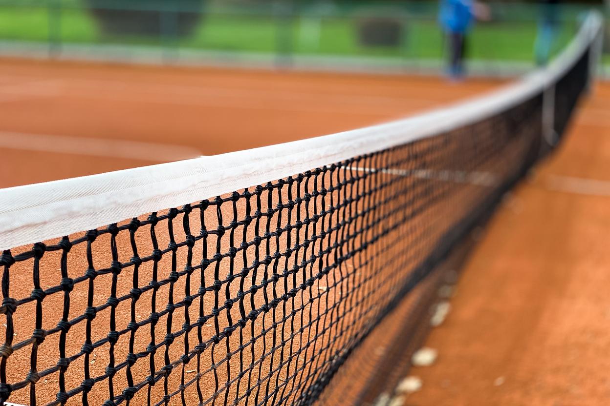 Nahaufnahme eines Tennisnetzes auf einem roten Sandplatz, Hintergrund unscharf mit grünen Bäumen.
